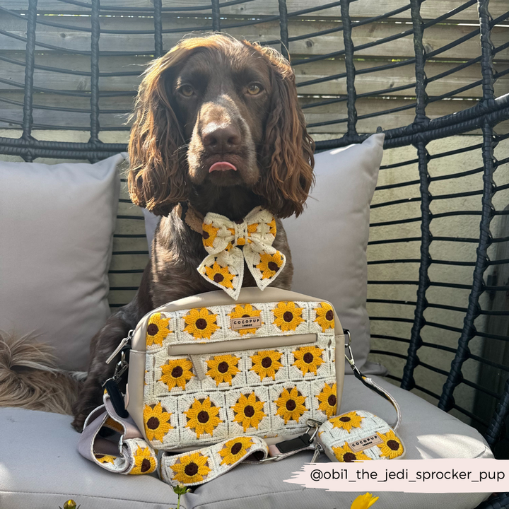 A brown dog with wavy ears sits on a swing chair. The dog wears a sunflower-patterned bow tie and is posed with a matching Dog Walking Bag featuring crochet-style fabric and Cocopup London's Bag Strap - Sunflower Patch. The background includes wooden fencing and outdoor cushions. Instagram handle @obil_the_jedi_sprocker_pup.