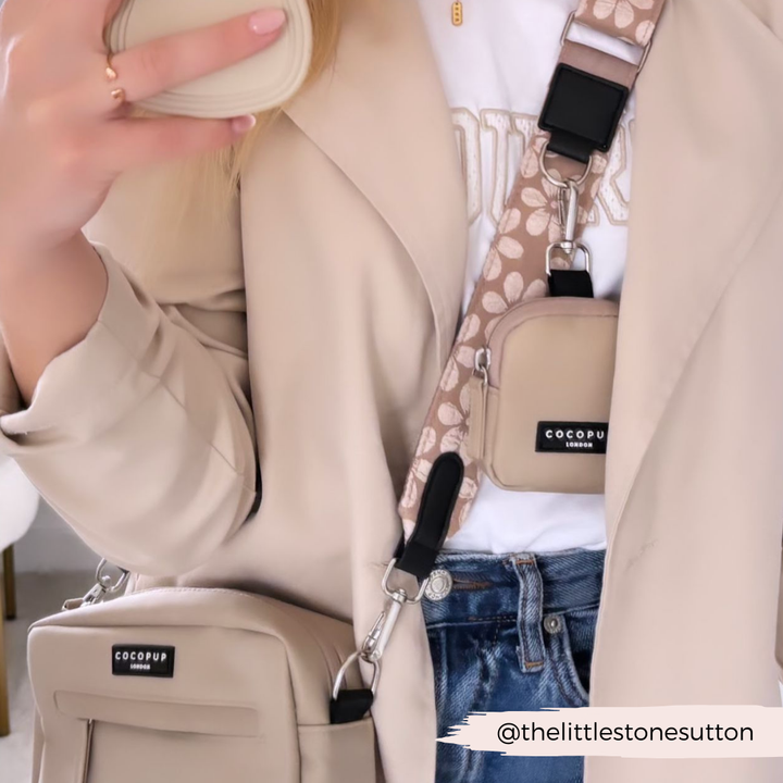 Close-up of a person wearing a beige coat, white top, and blue jeans. They're holding a beige smartphone. Two beige bags labeled "Cocopup London" are slung across their body. One bag features the Luxe Bag Strap - Mocha Flower in peach and white. The Instagram handle "@thelittlestonesutton" is displayed in the corner.