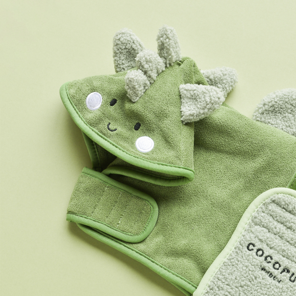 A Cocopup London PRE-ORDER Dinopup Drying Robe in green terry cloth, featuring an adorable dinosaur-themed hood with sewn-on eyes, a smiling face, and soft spikes on top. The premium microfiber towel is laid out flat against a light green background.