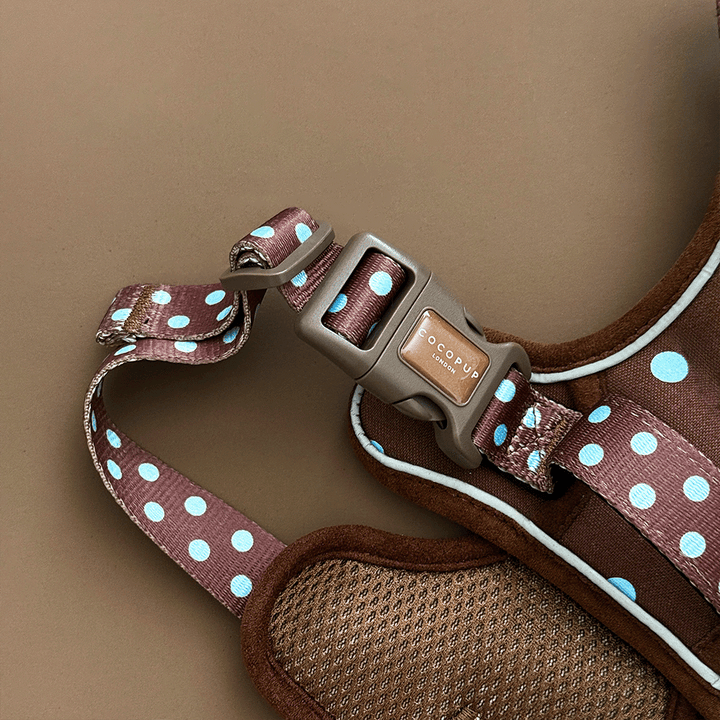 A close up of the buckle on a brown dog harness with light blue polka dots, laid flat on a solid brown background. 