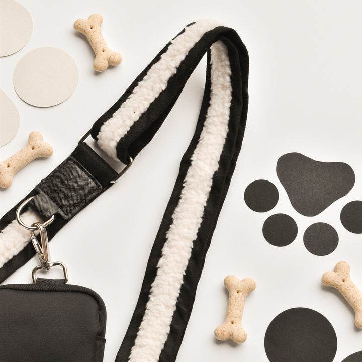 A black and white dog leash with a clip, a small dog treat bag, bone-shaped dog biscuits, and black and gray round cutouts alongside black paw print cutouts are arranged on a light surface. The faux suede Black Aviator Bag Strap by Cocopup London adds a touch of style, suggesting an elegant focus on dog walking and training.