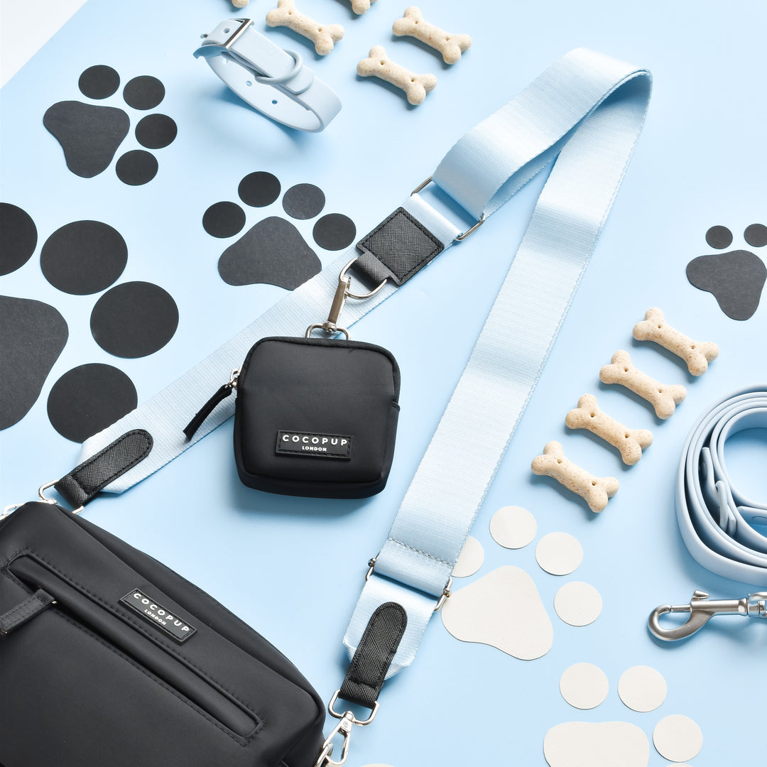 A neatly arranged display of dog accessories on a light blue background. Items include black pouches with Cocopup London's light blue NAKD Bag Strap in Bubblegum, a matching light blue leash, a white collar, several dog bone-shaped treats, and black and white paw print cutouts. The Cocopup Dog Walking Bag completes the stylish collection.