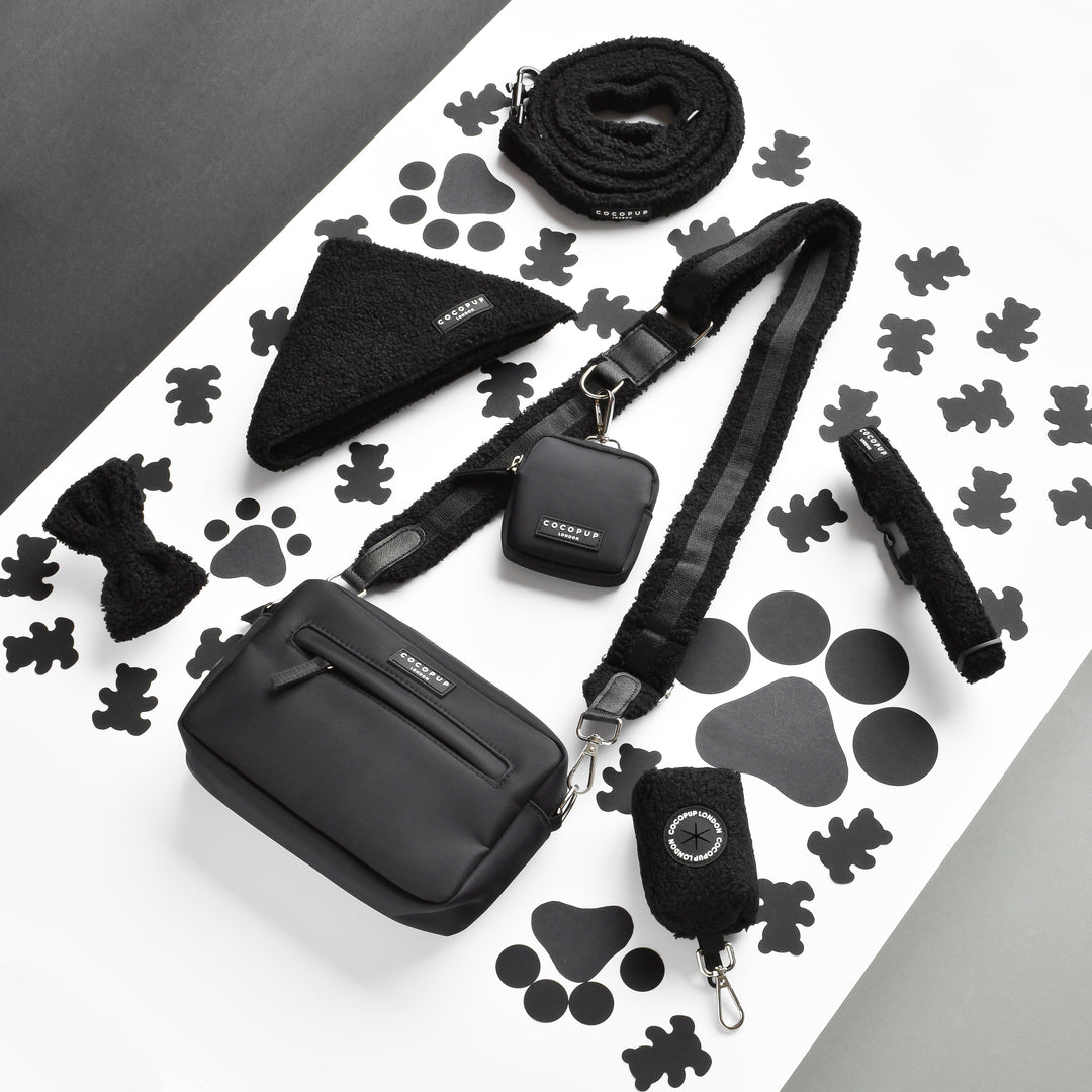 A sleek black pet accessory set is displayed on a white surface with black paw prints. The set includes a leash, a small pouch, a Cocopup London Dog Walking Bag with the Teddy Bag Strap - Baloo for that fluffy design, a bandana, a bow tie, and a poop bag holder—all matching in black fabric and detailing.