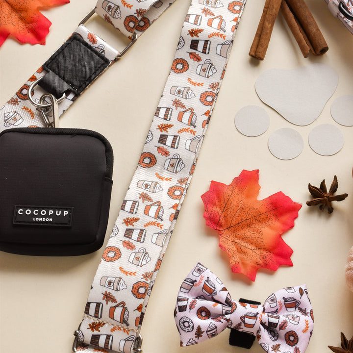 A "Bag Strap - Autumn Puppuccino" by Cocopup London, along with a white lanyard featuring a coffee-themed pattern, a black pouch labeled "Cocopup London," and a matching bow tie, are laid out on a beige surface. Surrounding items include a red maple leaf, cinnamon sticks, star anise, circular paper cutouts—creating the perfect autumnal dog walking accessory set for the season.