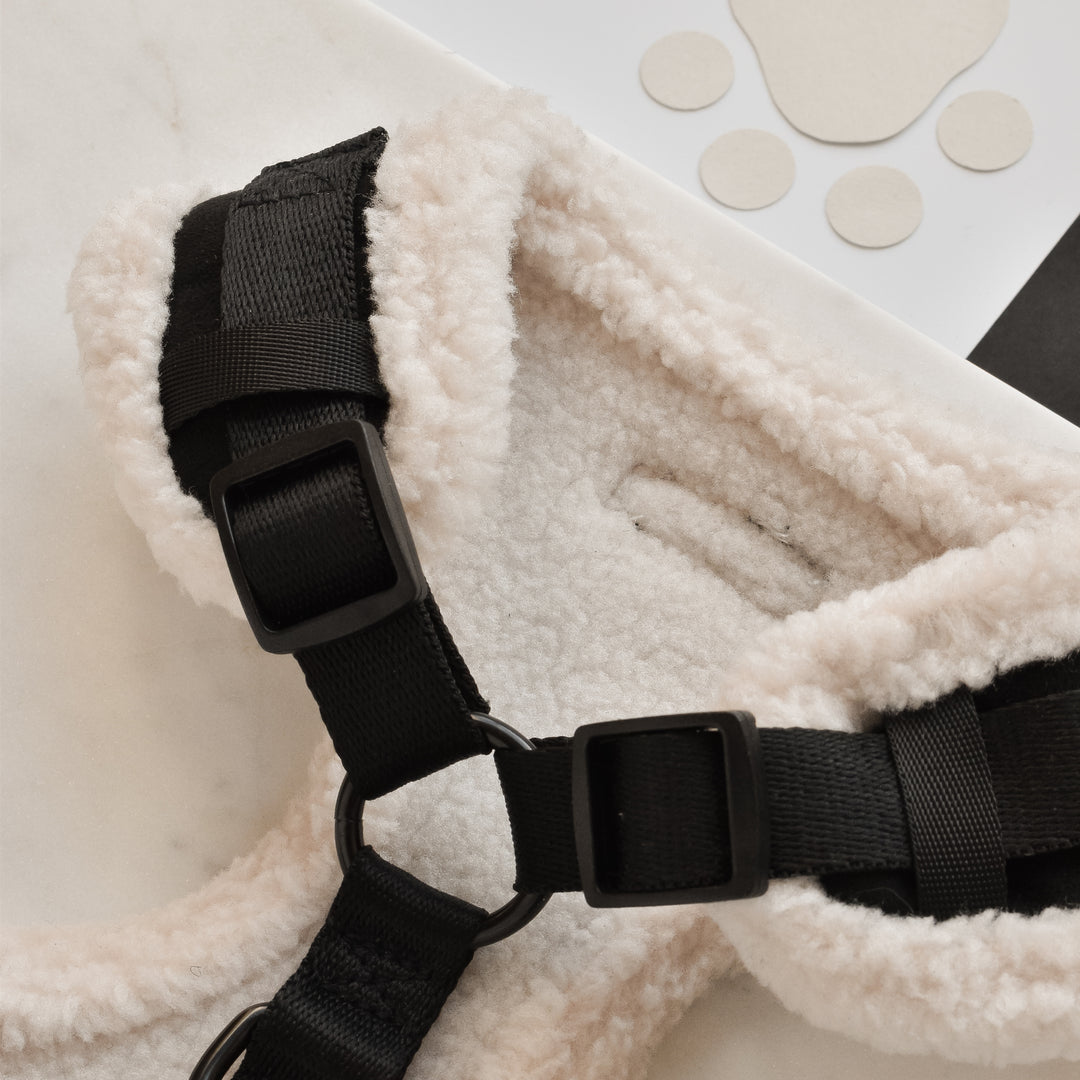 Close-up of the cozy, fleece-lined Black Aviator Adjustable Neck Harness from Cocopup London, featuring sturdy black straps and buckles. The harness sits on a marble surface, with a paw-print sticker and white sheet partially visible in the background.