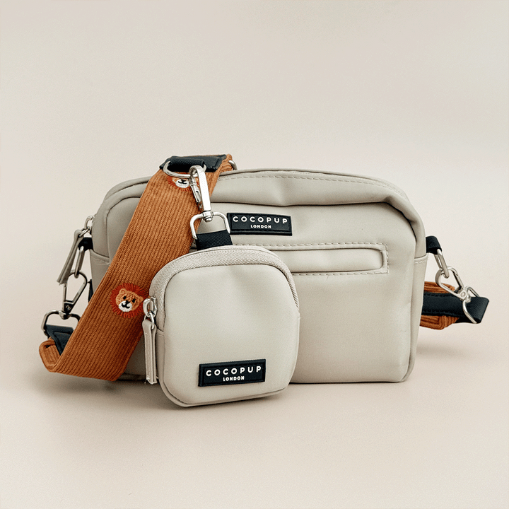 A light beige Cocopup Dog Walking Bag with a matching small treat pouch and a tan colour strap with lion embroidery, photographed on a plain warm beige studio background.