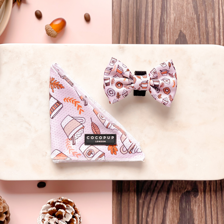 The Autumn Puppuccino Bandana and matching bow tie from Cocopup London, featuring a coffee and autumn-themed print, are displayed on a marble surface. Surrounding the items are pinecones, an acorn, and a textured wooden backdrop. The bandana includes a "COCOPUP LONDON" label, making it the perfect accessory for any coffee-loving dog.