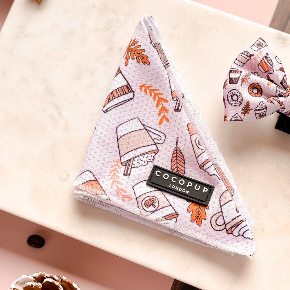 An Autumn Puppuccino Bandana with a light pink print featuring cups, leaves, and pumpkins rests on a marble surface. This bandana, part of the Cocopup London range of Bandanas, is perfect for the coffee-loving dog. A matching bow tie with the same pattern is partly visible next to it.