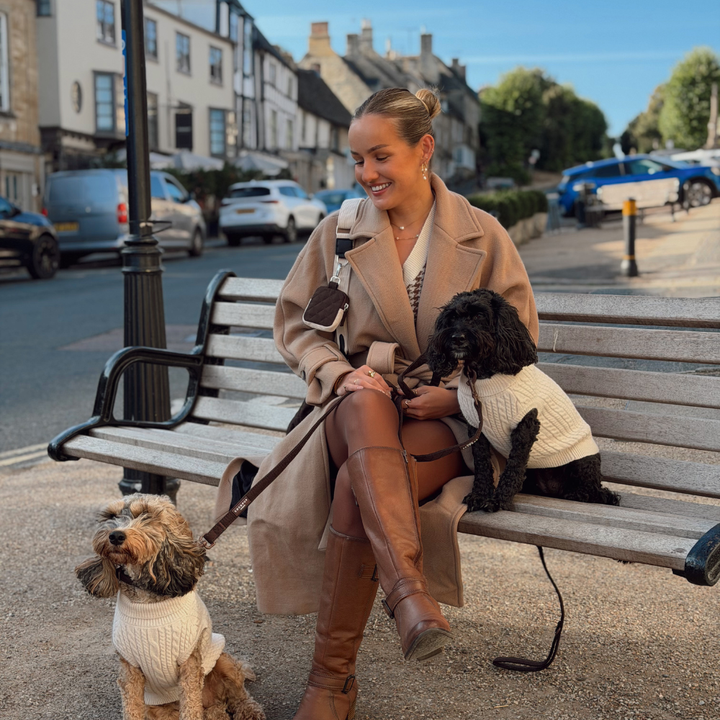 A woman sits on a wooden park bench outdoors with two dogs wearing cream knit sweaters. She is dressed in a camel coat, brown knee-high boots, and a white top, with a black bag slung over her shoulder.