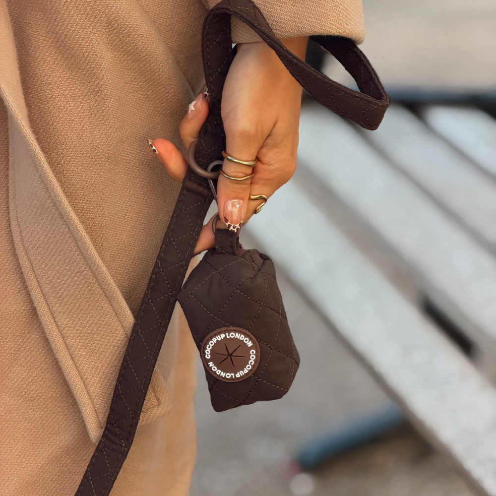 A close-up of a person holding a small brown quilted poop bag holder with a matching lead. 