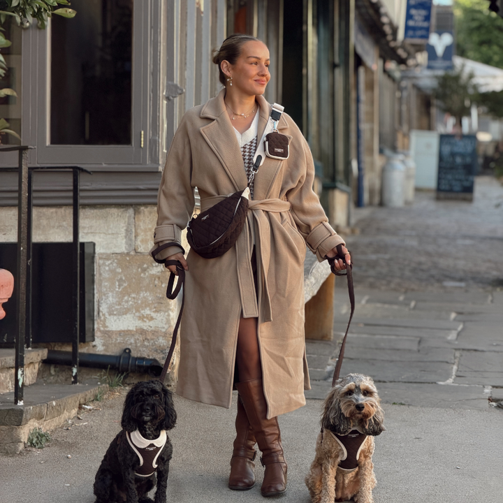 A woman walks two dogs on a city street wearing a long beige trench coat, brown knee-high boots, and a dark brown crossbody bag.