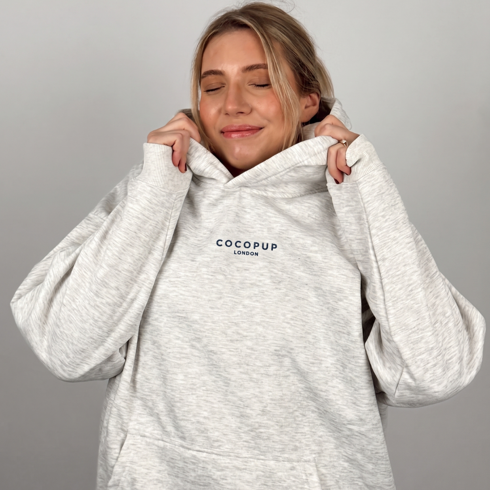 A woman wearing a light heather gray hoodie with 'COCOPUP LONDON' embroidered in black on the chest, posing with her hands on the hood against a plain gray background.