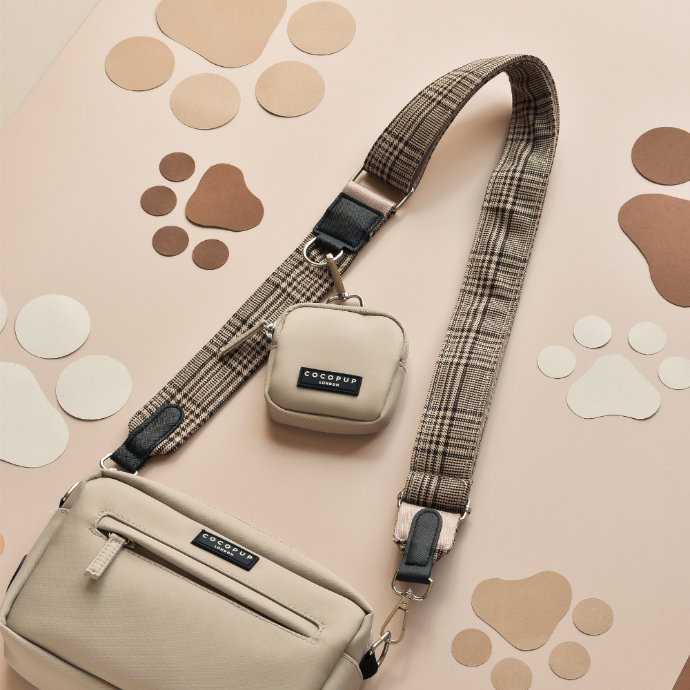 A beige Cocopup London Dog Walking Bag with a small matching pouch attached to its Bag Strap - Pup Plaid is placed on a light tan surface adorned with various brown and white paw print illustrations.