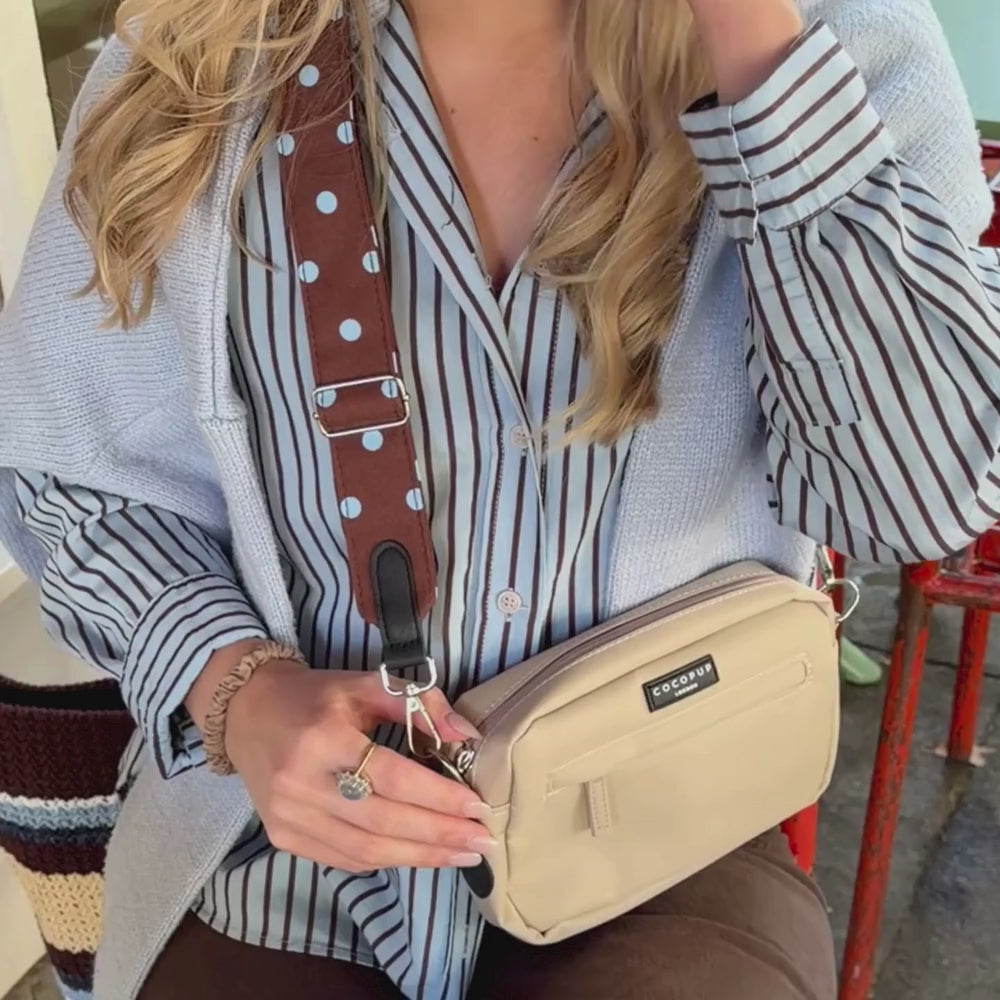 A woman wearing a light blue jumper over a blue and brown striped button-up shirt, holding a beige crossbody bag with a brown polka-dot strap. The bag has a black label that reads 'cocopup london'.
