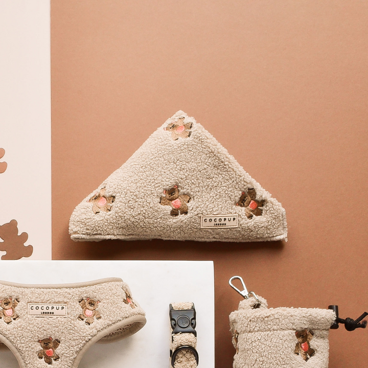 A set of dog accessories, including the Theo Embroidered Teddy Bandana, harness, and a bag adorned with a teddy bear pattern, are neatly displayed on a flat surface. These items are part of the Cocopup London range and feature the brand's label. The background consists of light peach, white, and brown hues.