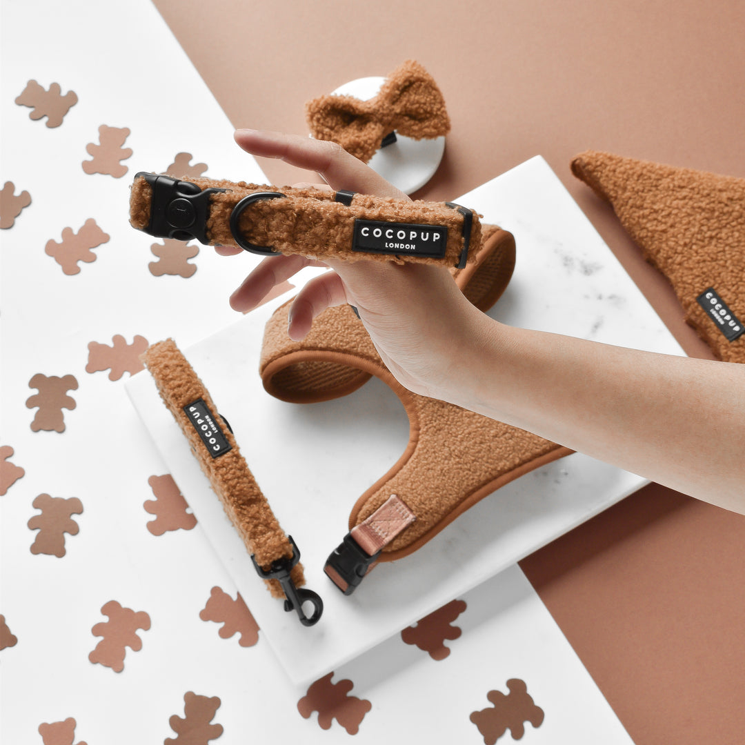 A hand holds a brown, fluffy dog collar featuring a black label with "Cocopup London." The same brand's Teddy Paddington Adjustable Neck Harness, Lead & Collar Bundle, which includes a neoprene harness, leash, and bow tie in brown tones, is displayed on a white marble surface surrounded by teddy bear-shaped cutouts.