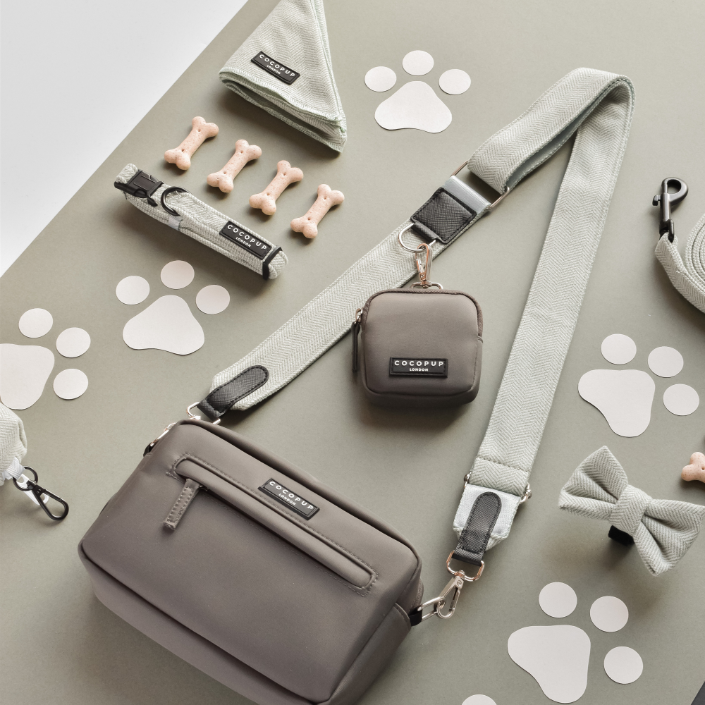 A flat lay image of pet accessories, including a grey crossbody bag with an adjustable Cocopup London Tweed Bag Strap - Sage, a smaller pouch, a leash, a collar, a bow tie, waste bag dispenser, dog treats shaped like bones, and paw print cut-outs on a grey background.