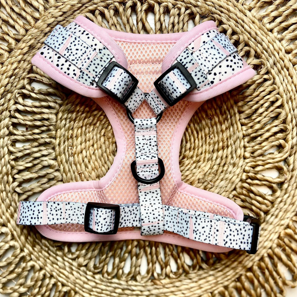 A Pink Dalmatian Adjustable Neck Harness from Cocopup London with white straps featuring black speckles and sleek black buckles lays on a woven mat. This stylish alternative to collars showcases multiple adjustable buckles and a metal D-ring for leash attachment, making it the perfect dog walk accessory.